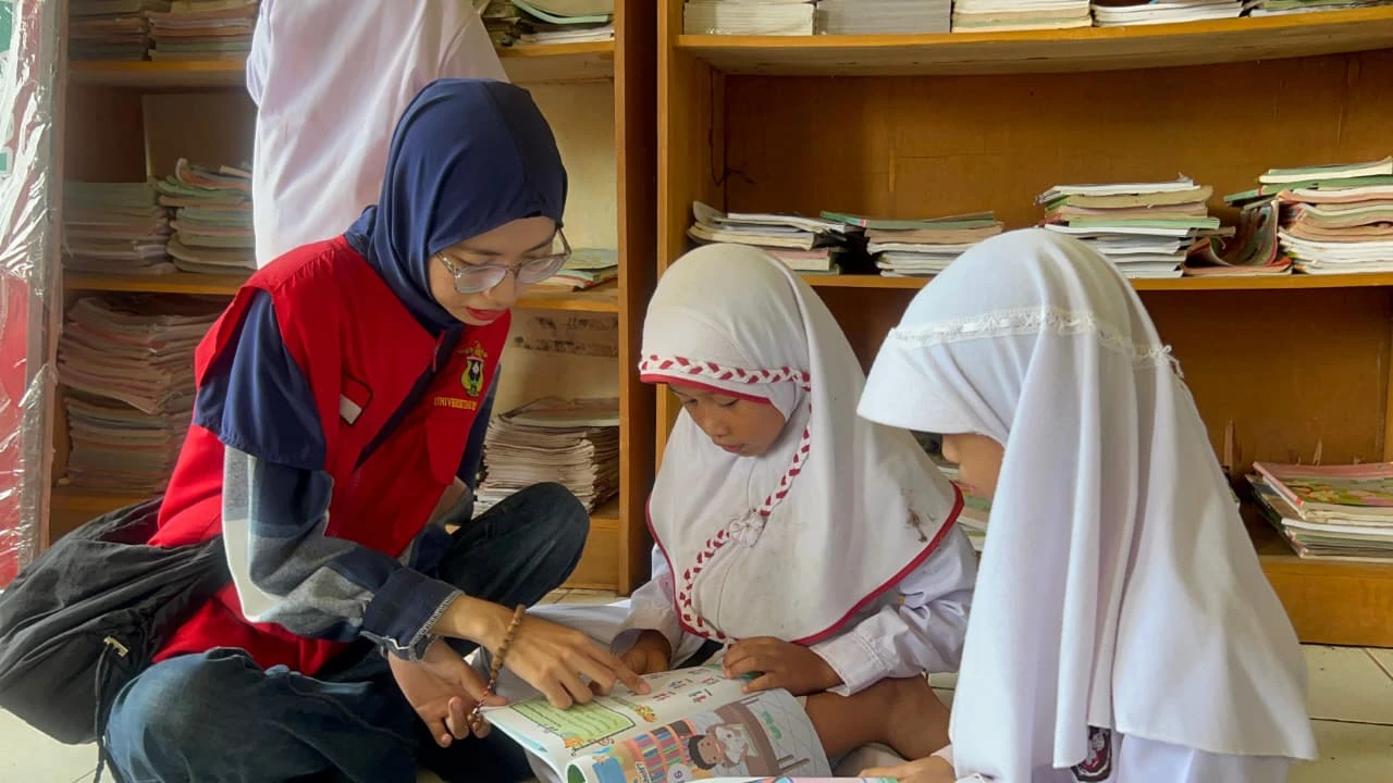 Sesi 'Membaca Bersama' yang menjadi bagian inti dari program KKN Tematik Literasi UNHAS. Seorang mahasiswa dengan sabar membimbing dan mengajak dua siswi SD Inpres Passuakkang untuk menyelami cerita dari sebuah buku. Momen ini bukan sekadar kegiatan, melainkan upaya menciptakan ruang nyaman agar anak-anak dapat berinteraksi dengan buku, menumbuhkan kecintaan pada literasi yang akan menjadi bekal masa depan. Kredit: Mahasiswa KKN Unhas.